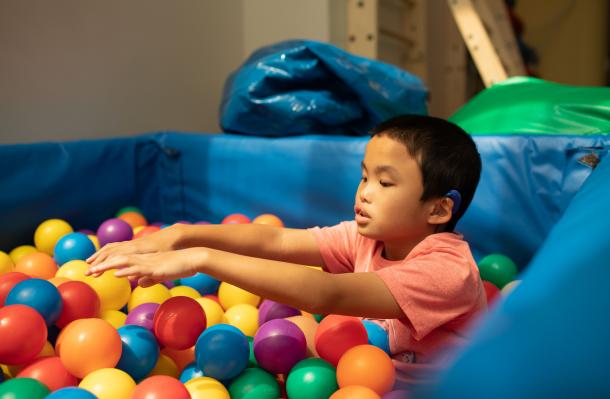 Manhattan Star Academy student is in a ball pit, their arms are outstretched, they wear a peach color shirt and hearing aid
