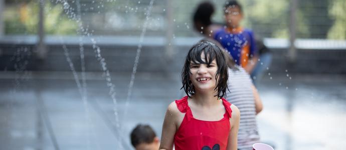 Manhattan Star Academy student outside playing in water sprinkler, they wear a red bathing suit and holding a red cup