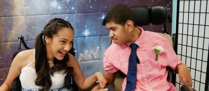 Jessica and Omar are both in wheelchairs right next to each other. It's prom and Omar is only looking at Jessica. The background is a large space/cosmic image.