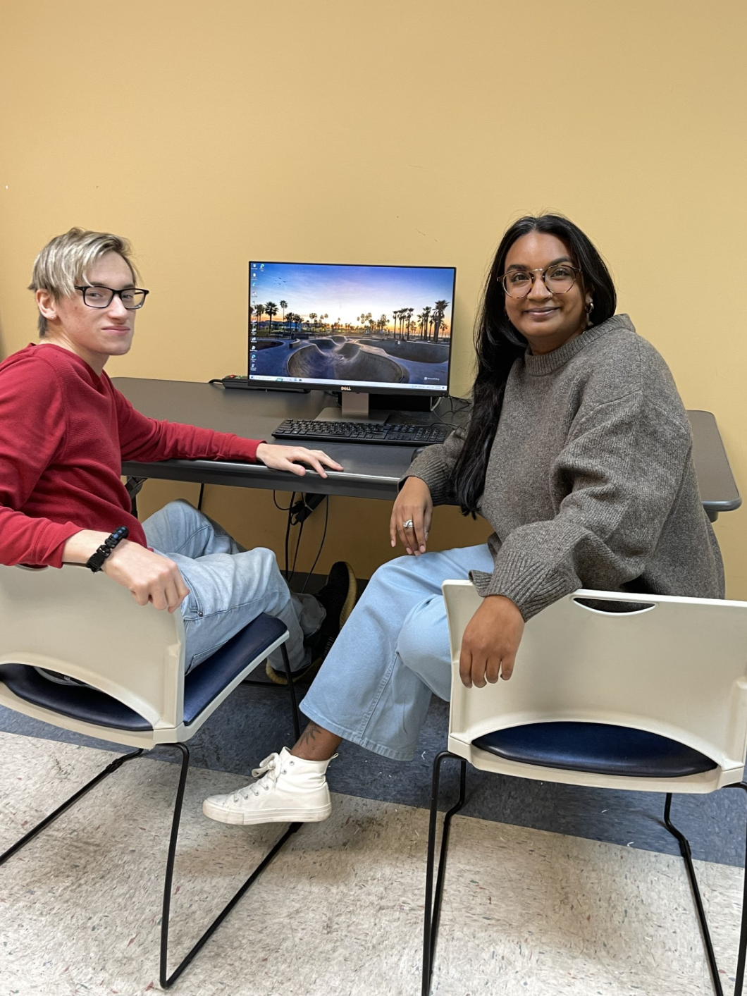 Two people sit on chairs at a desk with a computer, they are turning around to look at the camera for the picture