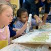 Kristina and Arthur are at a table, they are holding paint covered items with paint covered canvases in front of them.