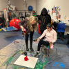 MSA student sits and looks on as a staff person leans over a canvas and looks to be pouring red paint