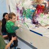 MSA student stands with support as they look on at a a colorful painting on a large sheet of paper on the wall