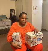 Latasha wears orange while sitting at a chair in a bedroom, she is holding boxes for small kitchen appliances