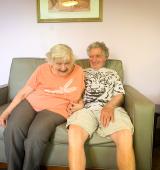 Joan and Melvin sat together on the couch, Joan is leaning forward and smiling, Melvin is looking at Joan