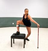 person in a leotard in a studio stands with one foot on a stool, they are holding a cane