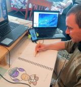 Sean sits at a desk, he has a computer on another level, but in front of him he has a sketchbook and is drawing in it