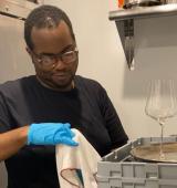 Jawann is standing in front of a fridge, drying a wine glass and a tray using a towel.