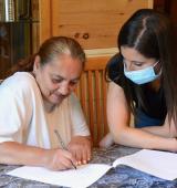 Two people sit at at table. One on the left is writing on a piece of paper, the other on the right is looking on and wears a mask.