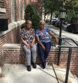 2 people sit on a wall outside an apartment building, they are posing for the photo
