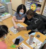 Three people sit at a table with a collection of artwork. They are actively engaged in the activity at hand (Luz Macias, Laurie Silver-Lewis, and Lavette Poinsette at work in the new Tarrytown Art Studio)
