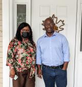 Two people stand in front of a door, one is wearing a flowery shirt and mask, the other is wearing a light blue button down shirt.