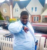 Person in blue button down shirt and tie stands outside looking at his watch. You see houses and cars in the background