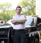 Person in black pants and white shirt leans against a black truck outside hospital (you can see an "emergency" sign in the background)