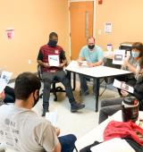 Group of people sitting in a circle in a room, they are holding paper or brochures in their hands and wearing masks