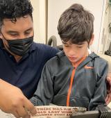 iHOPE student and staff sit together looking at a book, the student is in a wheelchair with a tray and you can see an attached communication device clamped in front of them