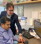 One person sits at a desk with keyboard and monitors in front of them, another leans over them to press keyboard