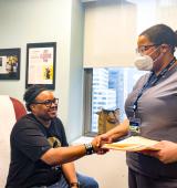 Darren sits in a doctors office and shakes hands with a person wearing a mask and scrubs, holding a manila folder