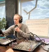 Two people sit at a table with mics and a mixing desk, they are wearing headphones and appear to be talking