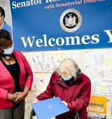 Two women stand in front of a wall with printed and hand made posters. One has a walker and holds a folder while the other looks at them. They both wear masks