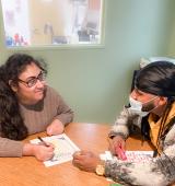 Two people sit at a desk, Karima on the left is writing, they are both looking at each other