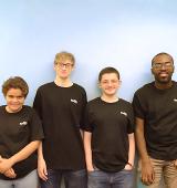 Group of 4 people stand leaning against a wall, all wearing black FedEx shirts and smiling for picture