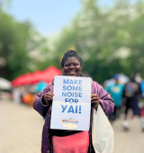 Destiny stands in Central Park holding a sign that says "Make some noise for YAI"