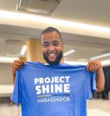 Breon smiles and poses for photo holding up a project SHINE t-shirt