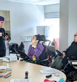 Group of people sit and stand around a table. They have AAC devices and snacks on the table