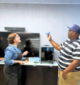 Two people stand in a room with various technological items. They are facing one another while the person on the right holds up a 3D printed item