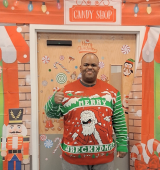 Kenneth stands and poses in his ugly Christmas sweater in front of a Christmas-themed backdrop
