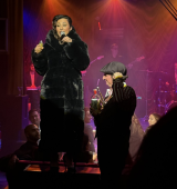 Angie Pontani in a dark fur coat sings into a microphone on a stage while another performer stands front; band members and drum equipment sit behind them under colorful spotlights.