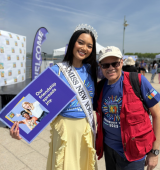 Two people smiling outdoors at an event, Miss New York is wearing a sash and a crown-like headpiece and holding a program booklet, standing beside Ismael in a cap and red vest on a sunny day.