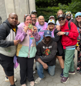 Group of Self Advocates posing outdoors by the bandshell in Central Park and wearing colorful clothing, with one person holding a small medal award.