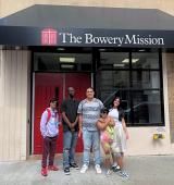 Group of people standing in front of a storefront entrance labeled “The Bowery Mission” with glass doors and brick walls on a New York City street.