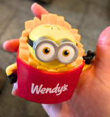Ashley's hand holds a small red and yellow “Wendy’s” minion toy (minion in fries with large googly-style glasses)