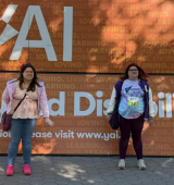 Ashley (right) and her sister stand in central park in front of a large orange sign with “YAI Seeing Beyond Disability,” text.