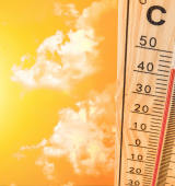Close-up of a mercury thermometer marked with C and F scales, with a red liquid line, set against a bright orange-yellow sky with scattered clouds.