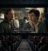 A packed movie theater watches a screen showing two men in a scene, with subtitles across the bottom that read “This is an example of open captions displayed on a movie theater screen”