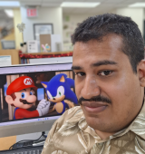 Orland sitting at a desk looking at a computer screen showing Mario and Sonic character artwork in an office.