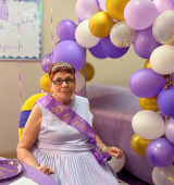 Veronica is seated indoors at her 80th birthday celebration. She is wearing a purple sash and glasses, with a large purple, white, and gold balloon cluster behind her and a hand-drawn “Happy Birthday” message on the wall.