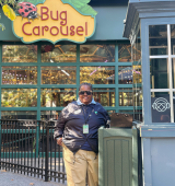 Person standing near a gray ticket booth in front of a colorful “Bug Carousel” sign, with a large glass building behind and autumn leaves framing the scene.