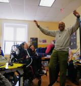 Man stands with arms in air in front of a smartboard and children and staff in classroom