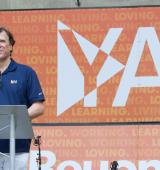 George Contos stands at a podium in front of a large YAI banner