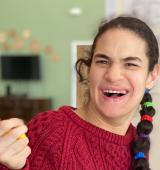 Heather wears a red sweater, has her right hand up, and is smiling