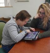 two women sit at a table, one is using an ipad while the other watches/supports