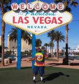 Omari wears a tie-dye shirt and stands in front of the welcome to Las Vegas sign