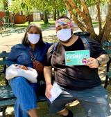 Two people wearing masks sit on a bench outside. One is holding a flyer about COVID testing