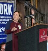 Woman standing at podium looking a little behind her. Banner behind her has the word "work" with YAI logo