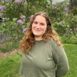 Person with long curly hair wearing a green top is stood outside in front of a lilac bush, they are holding their hands behind their back and is smiling